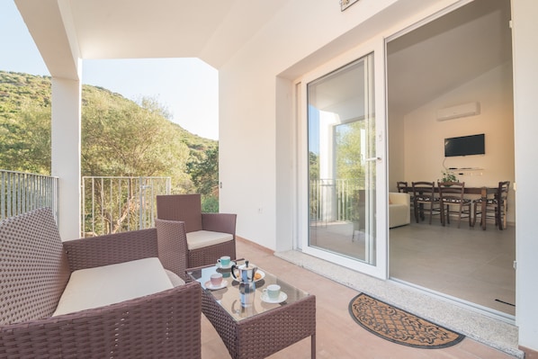 A balcony with wicker furniture, a glass table with cups and a coffee pot, and a sliding glass door leading to an indoor dining area with chairs and a TV.