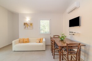 A modern living room with a sofa, dining table, chairs, a flat-screen TV, an air conditioner, and a window.