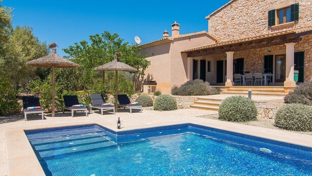 A pool with lounge chairs and umbrellas, a stone building with a covered patio, and a clear blue sky.