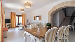 A dining area with a wooden table and white chairs, a sofa, a TV, and a staircase.