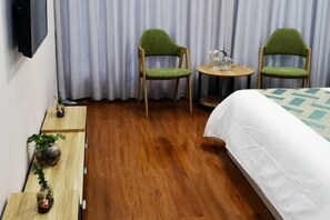 A hotel room with a wooden floor, a bed with a patterned bedspread, two green upholstered chairs, a small round table with a glass top, and a television mounted on the wall. - Kaibin Four Seasons Hotel (Renshou)