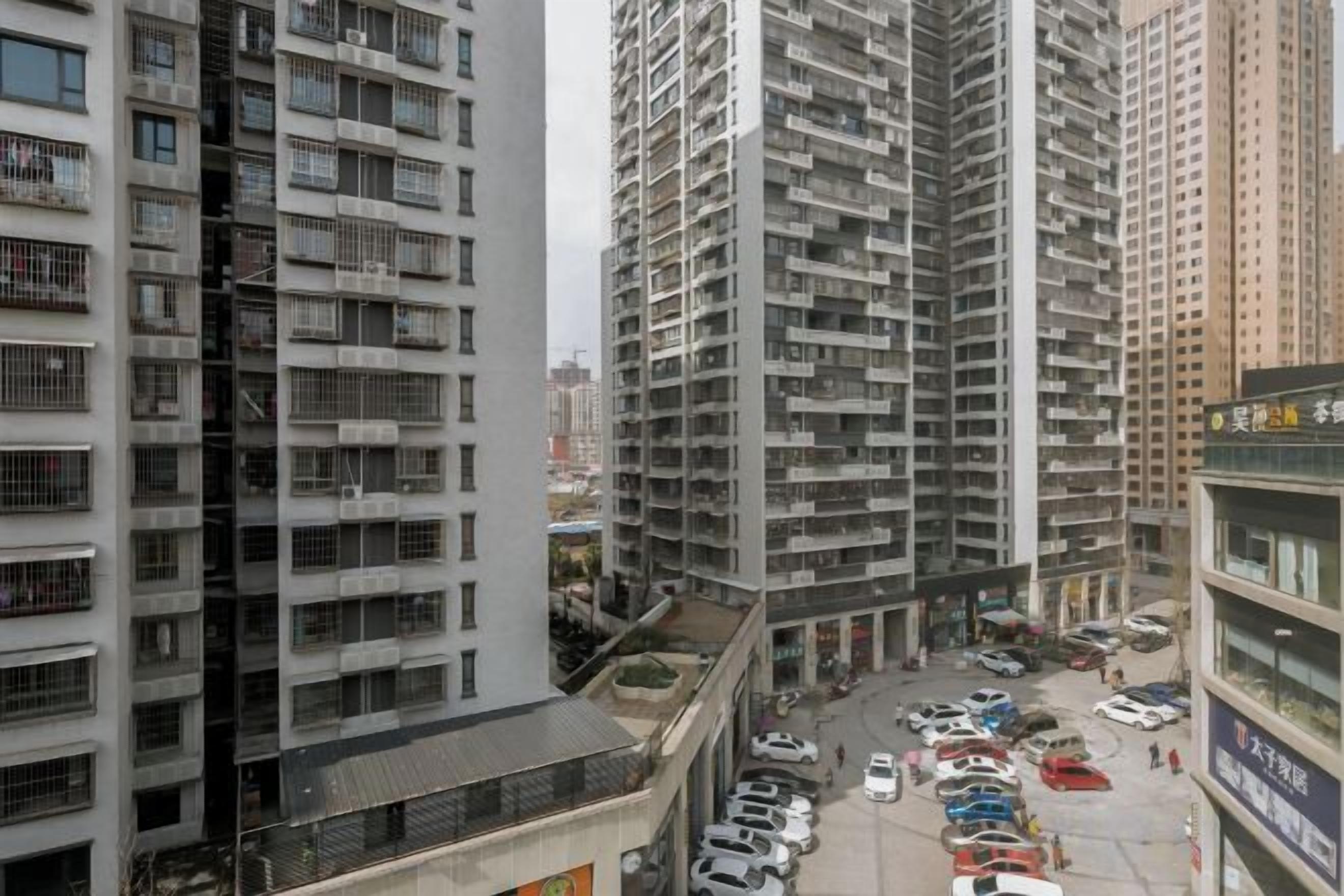 A high-rise residential building with multiple balconies, a parking lot with several cars, and a commercial building with signage.