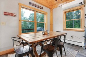 Una mesa de comedor de madera con cuatro sillas, dos ventanas grandes con vistas a árboles, un letrero que dice 'Monday Mourning' y un letrero de 'Maine Lobsters Vacationland'.