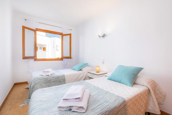 Ein Zimmer mit zwei Betten, einem Fenster mit Vorhängen, einem Nachttisch mit Lampe und einer Wandleuchte.