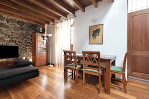 Una habitación con techo de madera, pared de piedra, televisor de pantalla plana, un sofá, una mesa de comedor con sillas y un cuadro en la pared.