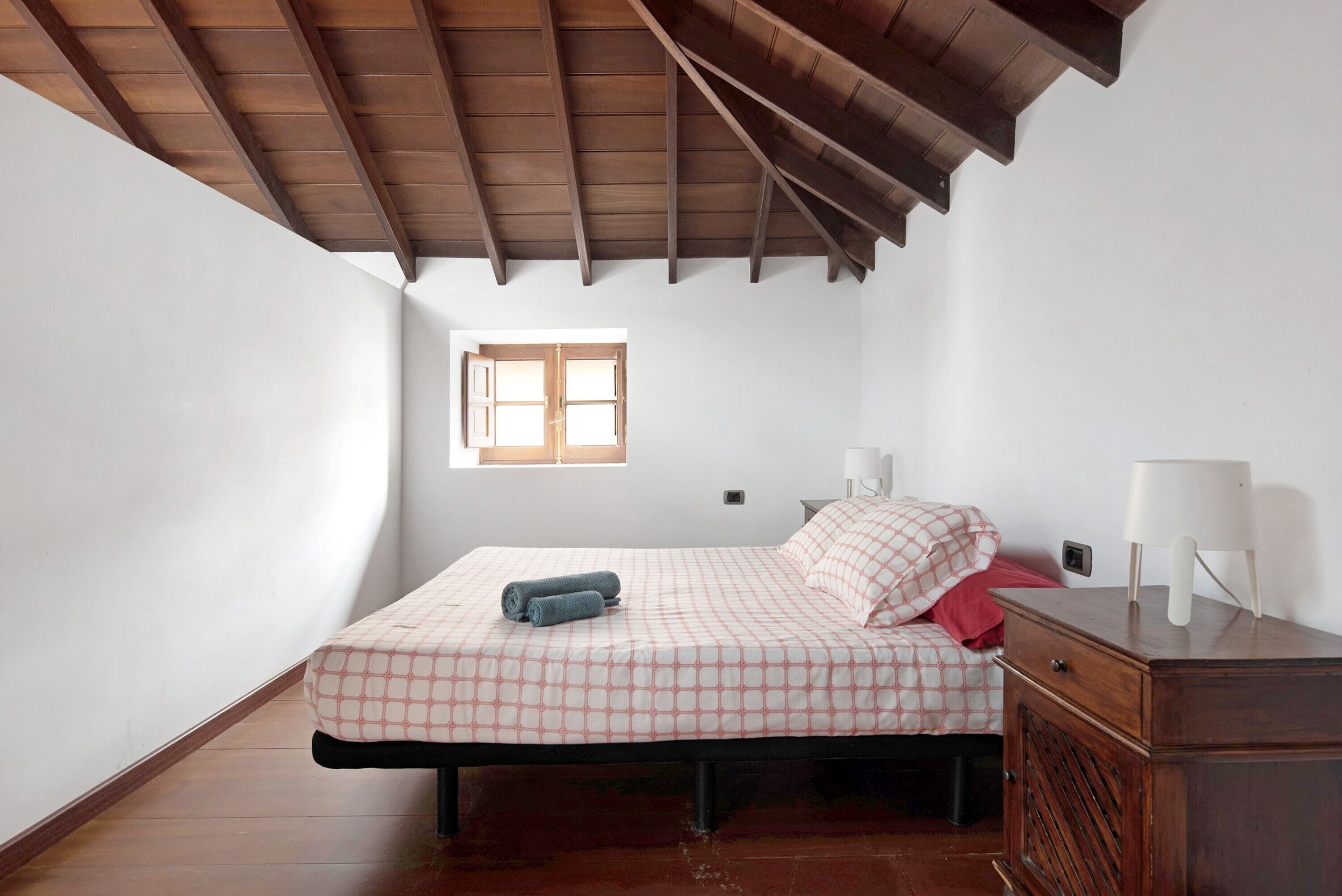 Un dormitorio con techo de madera, una cama con colcha a cuadros, una mesita de noche con lámpara y una ventana con persianas.