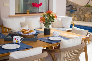 A dining area with a wooden table, wicker chairs, white cushions, a vase with flowers, and a basket of croissants.