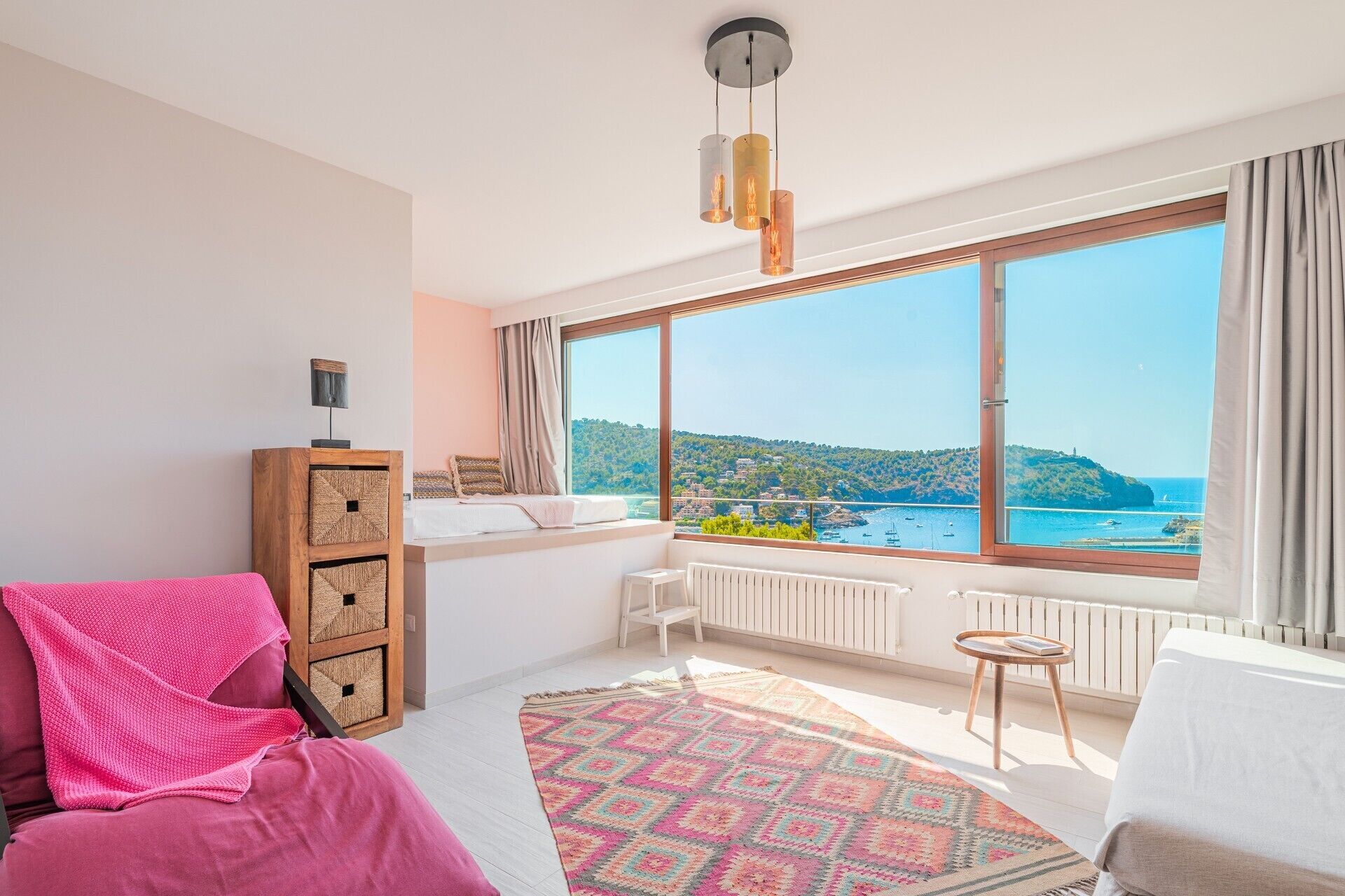 A bedroom with a large window, a bed, a bedside table, a chair, and a view of the sea.