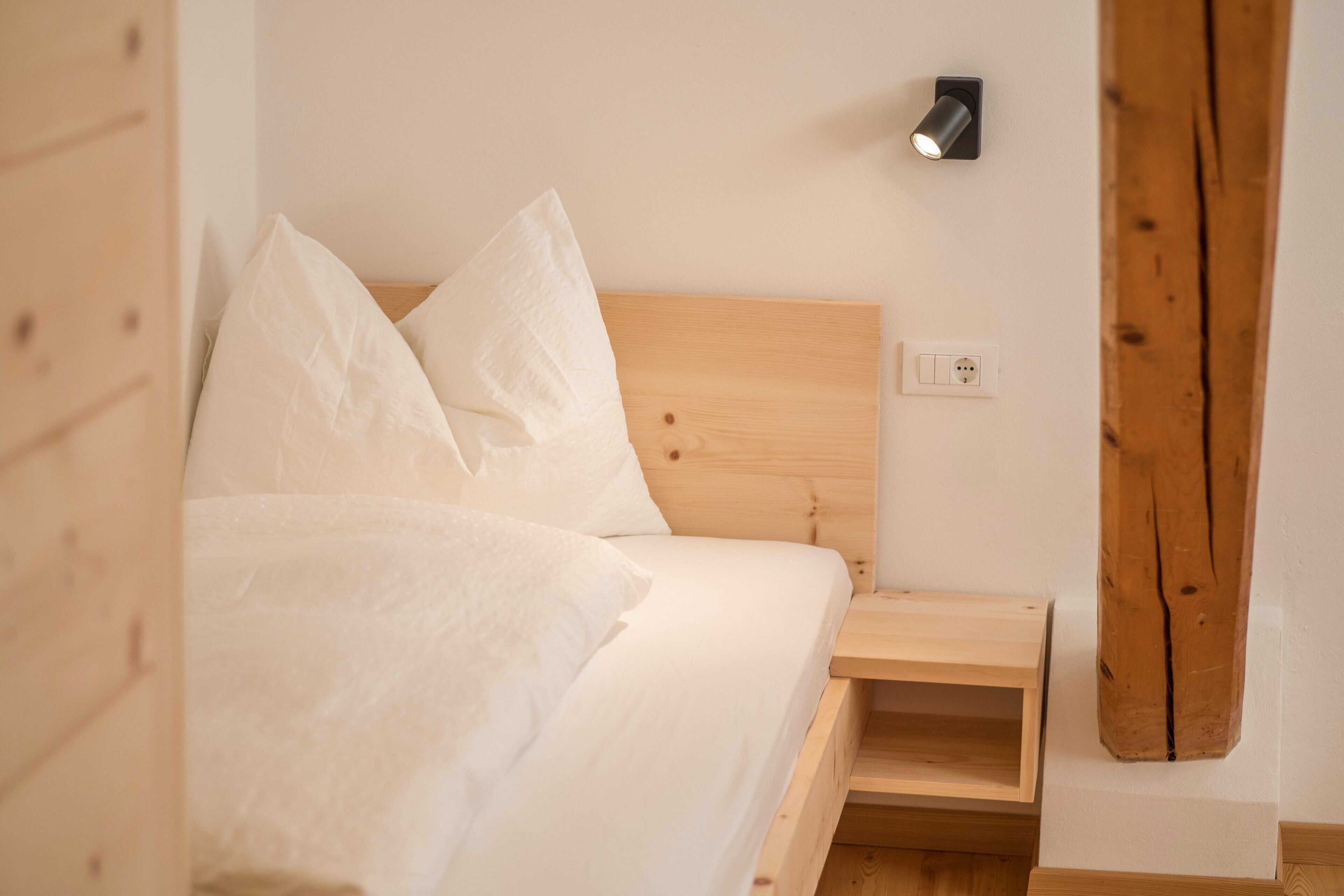 A bed with white linens, a wooden headboard, a bedside table, and a wall-mounted light fixture.