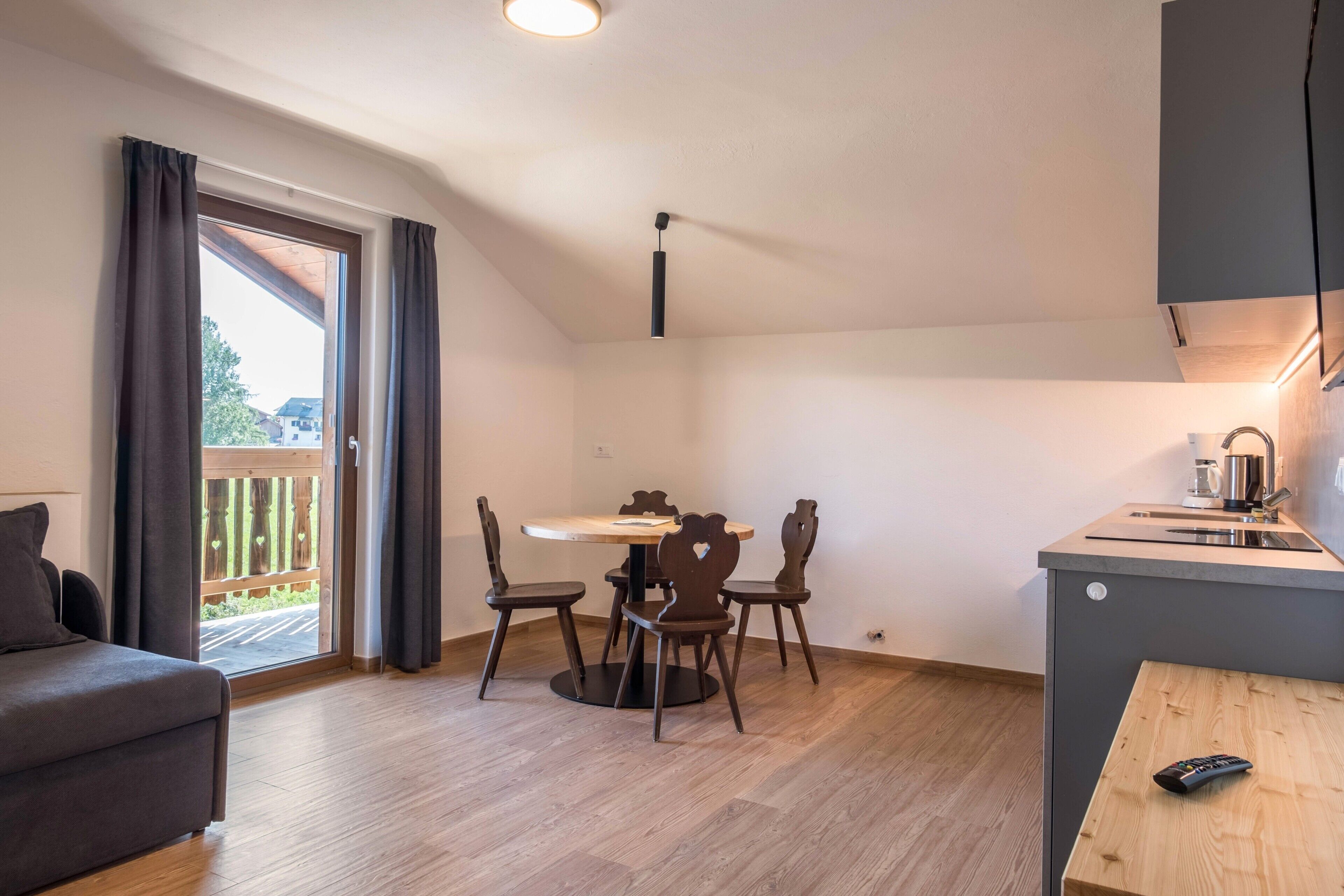 Un espace de vie compact comprenant une table à manger, quatre chaises, un canapé, un petit coin cuisine et un balcon avec des rideaux.