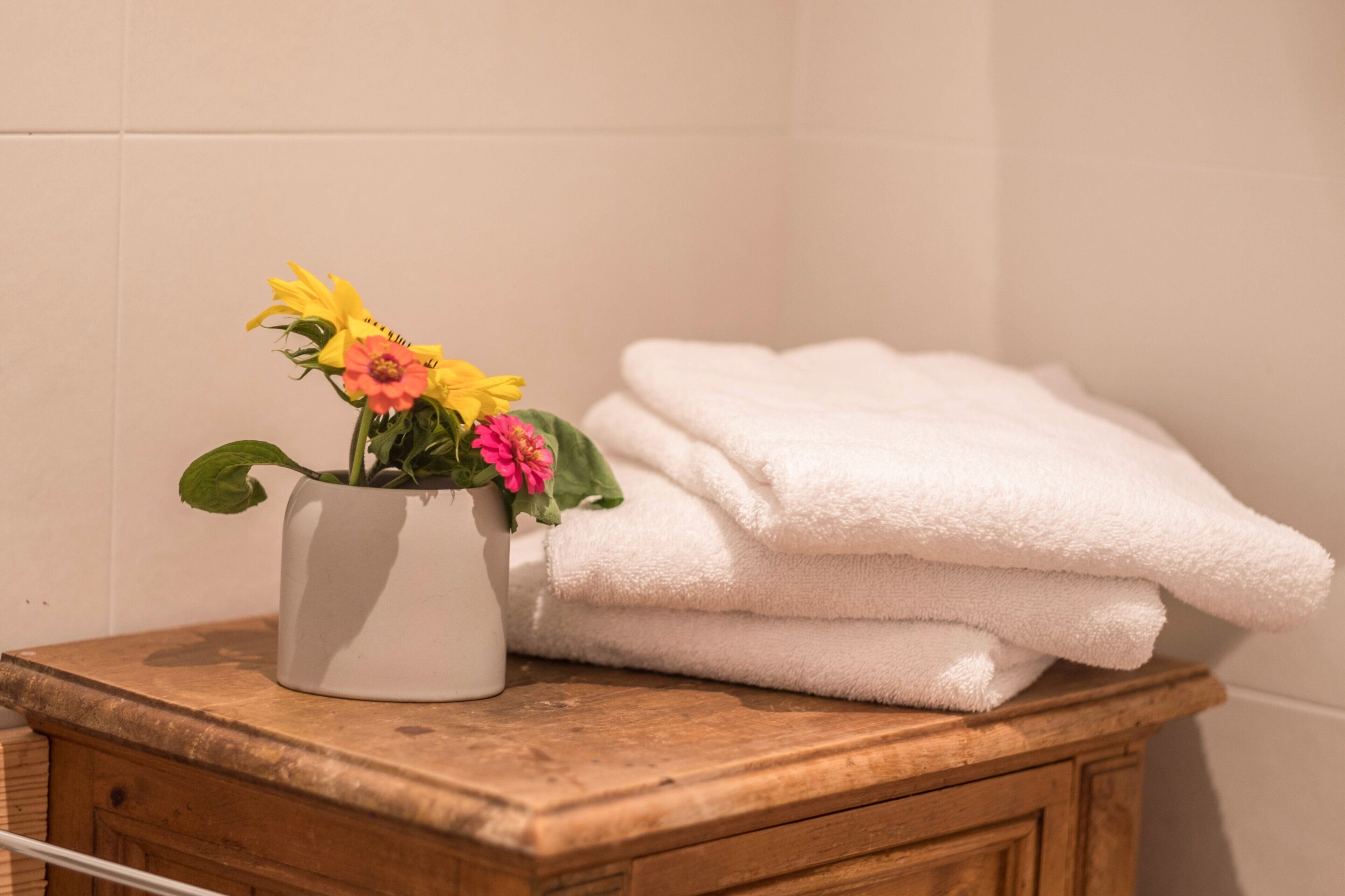 Ein Badezimmer mit einem Holz-Waschtisch, einer weißen Vase mit Blumen und gefalteten weißen Handtüchern.