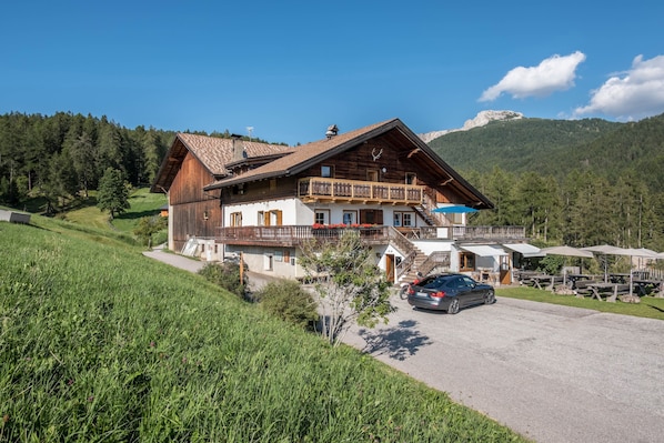 Un bâtiment en bois à deux étages, doté d’un balcon, avec une voiture garée devant et un paysage montagneux en arrière-plan.