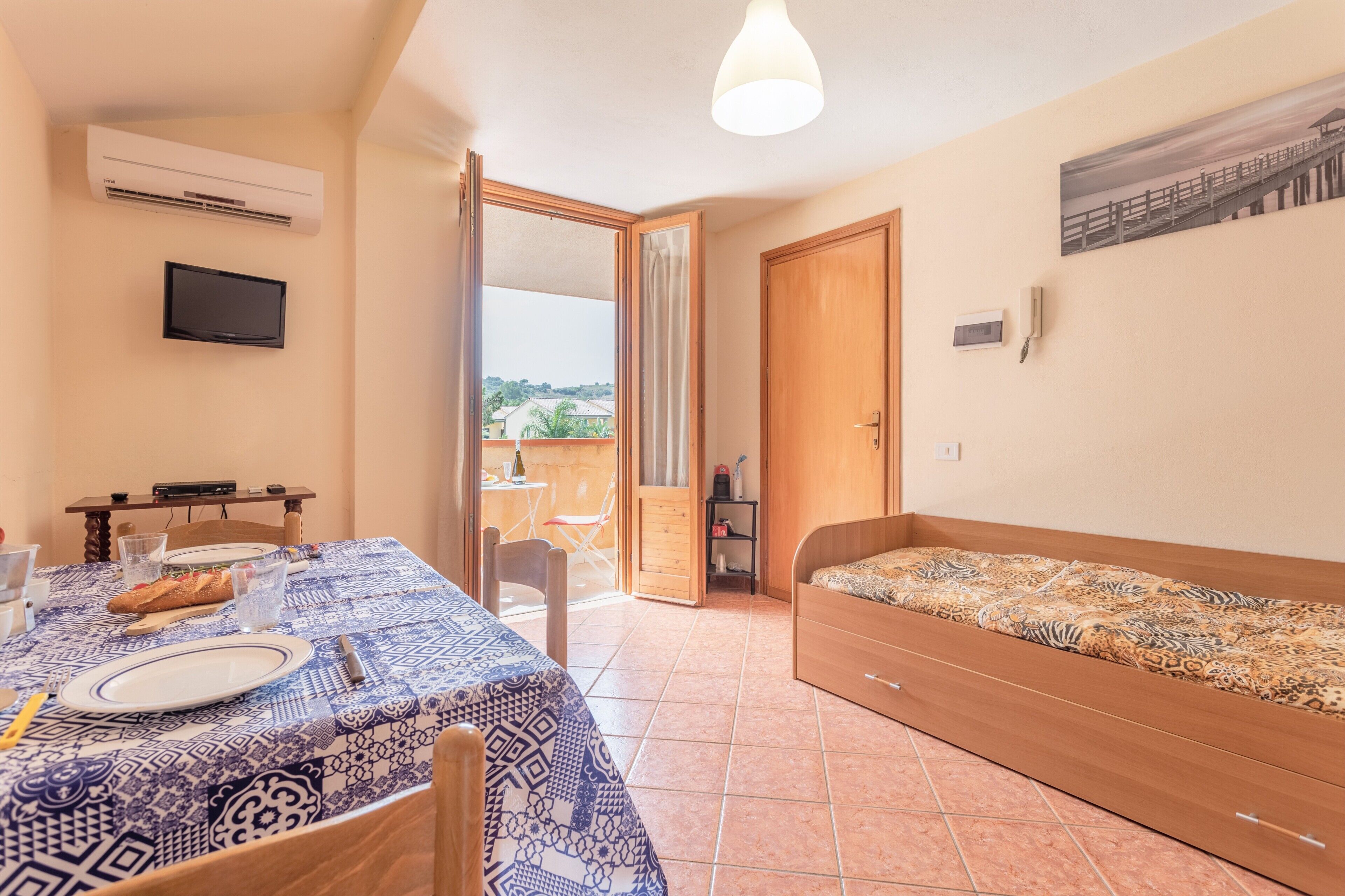 A room with a dining table set for two, a single bed, an air conditioner, a TV mounted on the wall, and a balcony with a view.