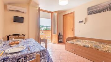 A room with a dining table set for two, a single bed, an air conditioner, a TV mounted on the wall, and a balcony with a view.