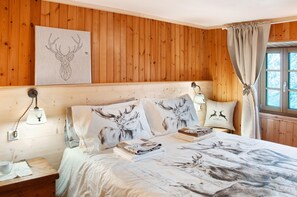 Une pièce aux murs lambrissés, avec un lit agrémenté d’une literie aux motifs de cerfs, une lampe fixée au mur et une fenêtre avec des rideaux.