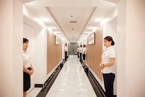 Um corredor de hotel com piso lustroso, quadros nas paredes e um funcionário uniformizado ao fundo.