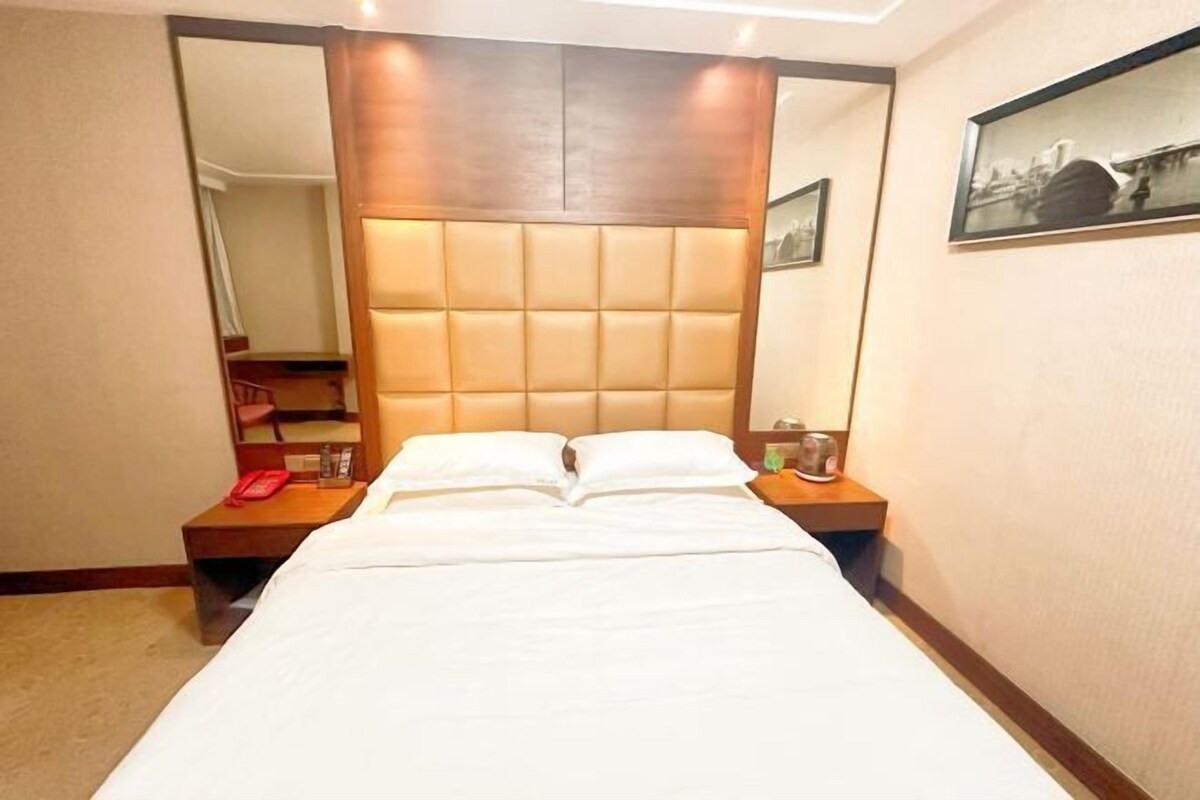 A hotel room with a large bed, a wooden headboard, a bedside table with a lamp, and a framed picture on the wall.