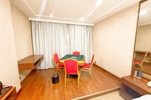 Una habitación de hotel con una mesa de billar verde, sillas rojas, suelo de madera, un cubo de basura, un escritorio pequeño con un espejo y una cama con ropa blanca.
