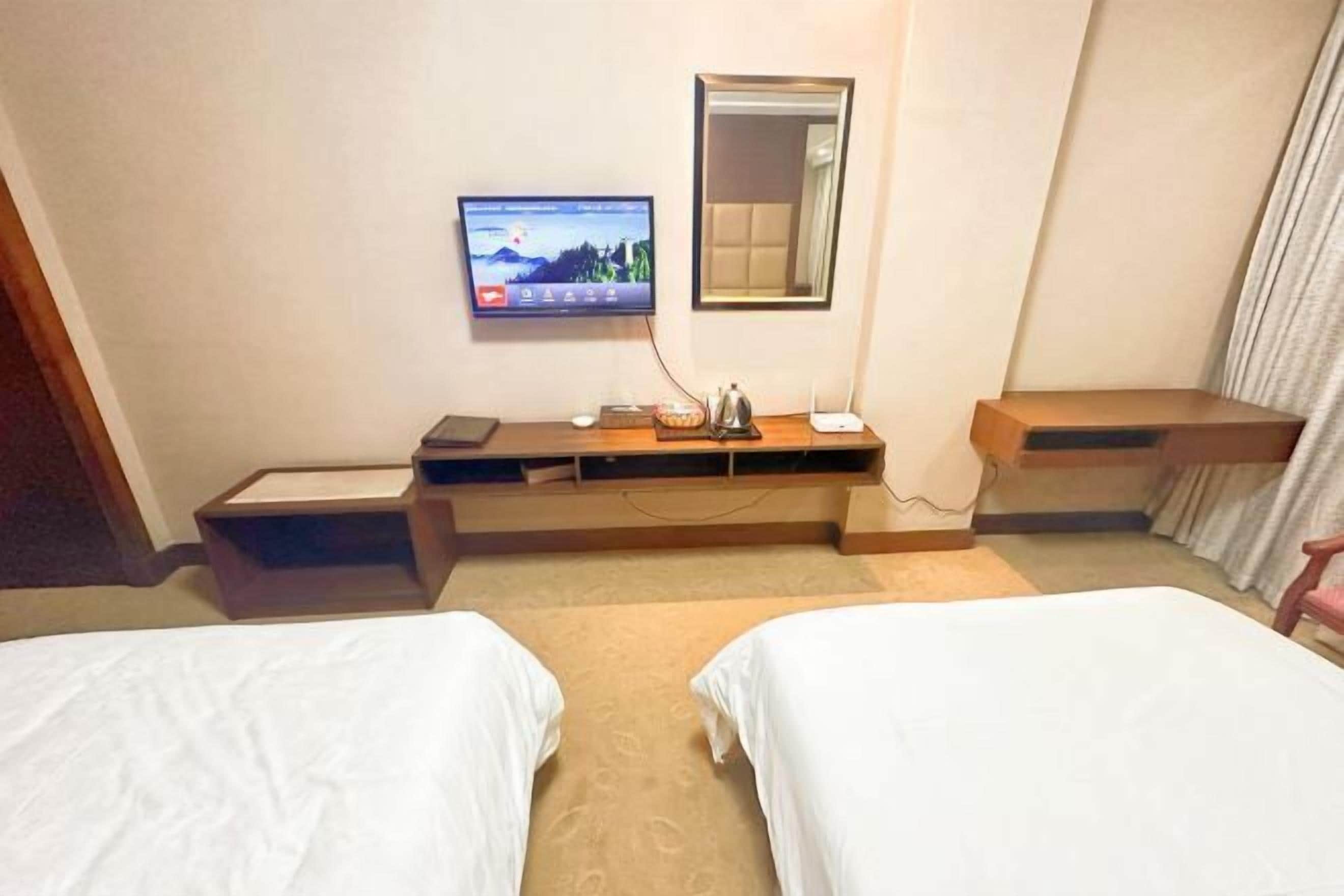 A hotel room with two beds, a TV mounted on the wall, a bedside table with a lamp, and a mirror above the table.