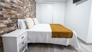 Een slaapkamer met een bakstenen muur, een bed met witte beddengoed en een bruine plaid, een witte nachtkastje met lades, en een aan de muur bevestigde televisie.