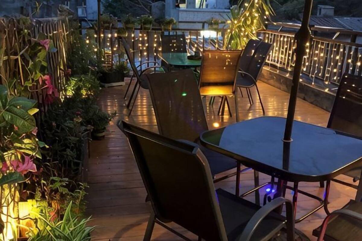 Un patio exterior con mesas y sillas, rodeado de plantas y luces colgantes.