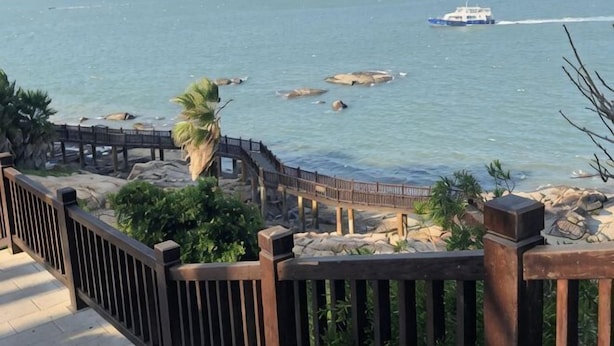 Vista costera con un paseo de madera, costa rocosa y un barco en el agua.
