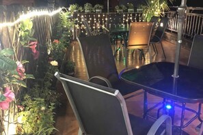 Un área de estar al aire libre con mesas y sillas, rodeada de plantas y luces colgantes.