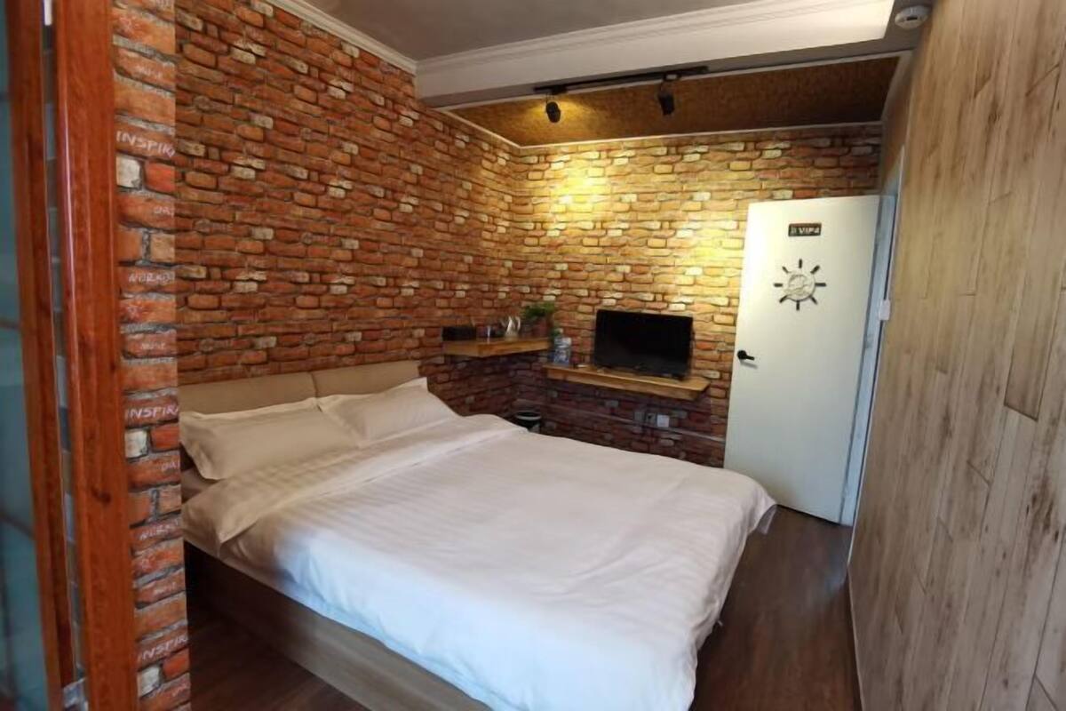 Un dormitorio con pared de ladrillo, una cama, un televisor y una puerta.