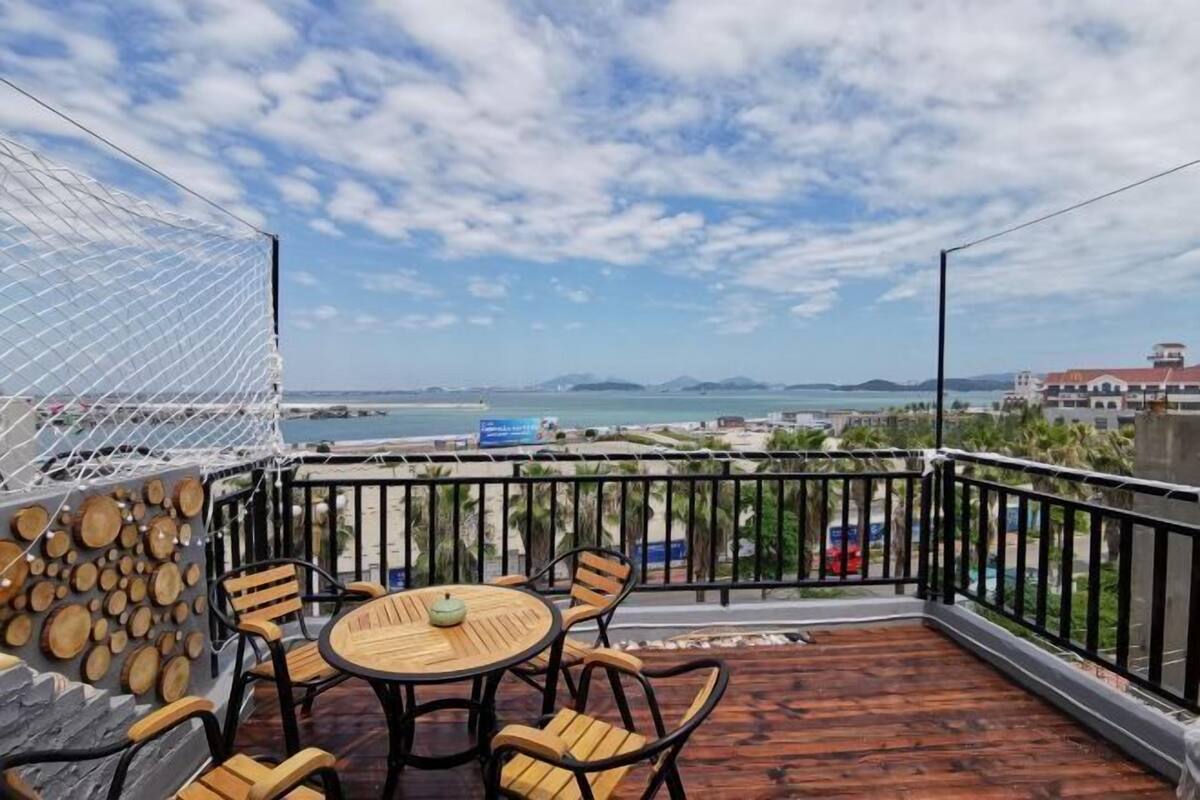 Una terraza de madera con una mesa redonda y sillas, una zona con mosquitero blanco y vistas al mar y edificios lejanos.