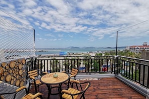 Una terraza de madera con una mesa redonda y sillas, una zona con mosquitero blanco y vistas al mar y edificios lejanos.