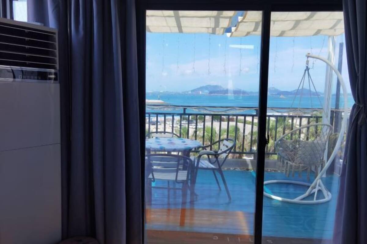 Un balcón con mesa y sillas, una silla colgante y vistas al mar y a la costa lejana.