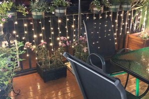 Un espacio de patio con una mesa de vidrio, sillas, plantas en macetas y luces colgantes.
