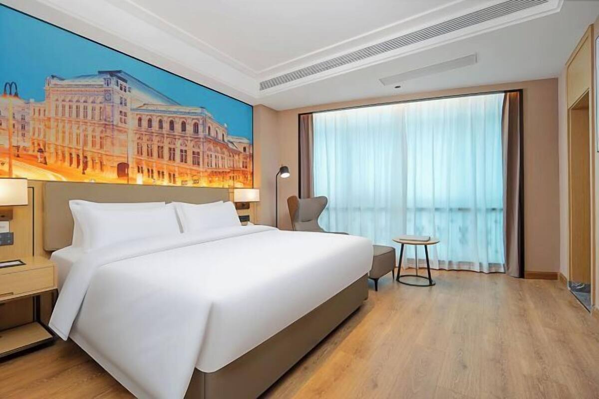 Une chambre d’hôtel avec un grand lit, une table de chevet, une lampe, une petite table et une grande fenêtre avec des rideaux.