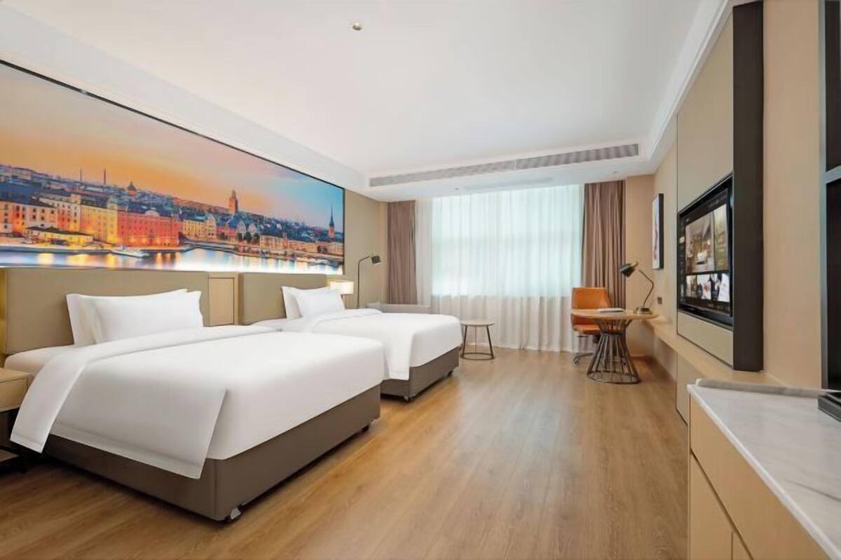 Une chambre d’hôtel avec deux lits, un bureau, une chaise, une télévision et un tableau représentant un paysage urbain.