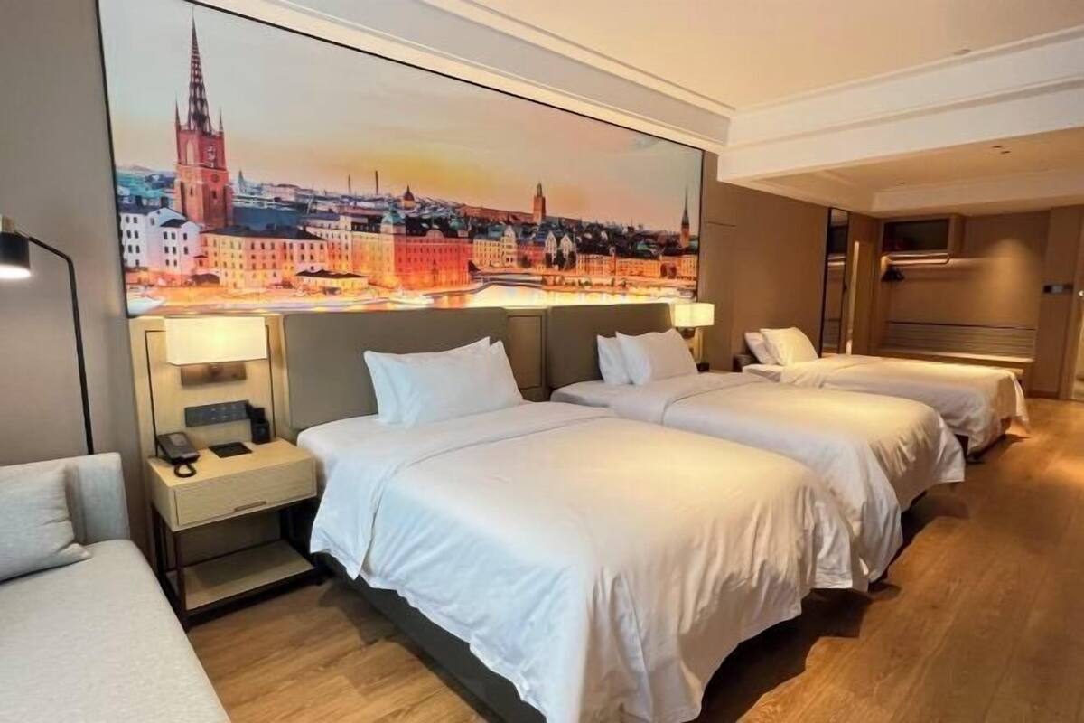 Une chambre d’hôtel avec deux lits, une table de chevet, une lampe et un grand tableau représentant un paysage urbain accroché au mur.