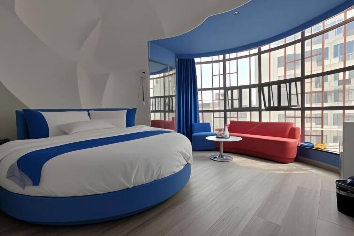 Una habitación de hotel moderna con una cama curvada, un sofá rojo, una mesita redonda y grandes ventanales con vistas a la ciudad.