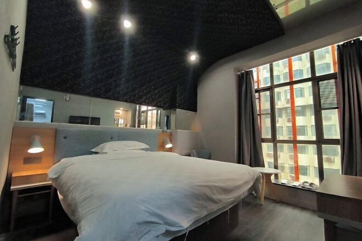 Ein Hotelzimmer mit Bett, Nachttischlampen, einem kleinen Tisch, einem Spiegel und einem Fenster mit Vorhängen.