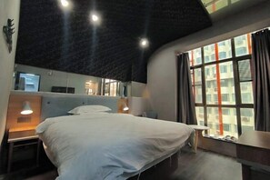 Et hotelværelse med seng, sengeborde, et lille bord, et spejl og et vindue med gardiner.
