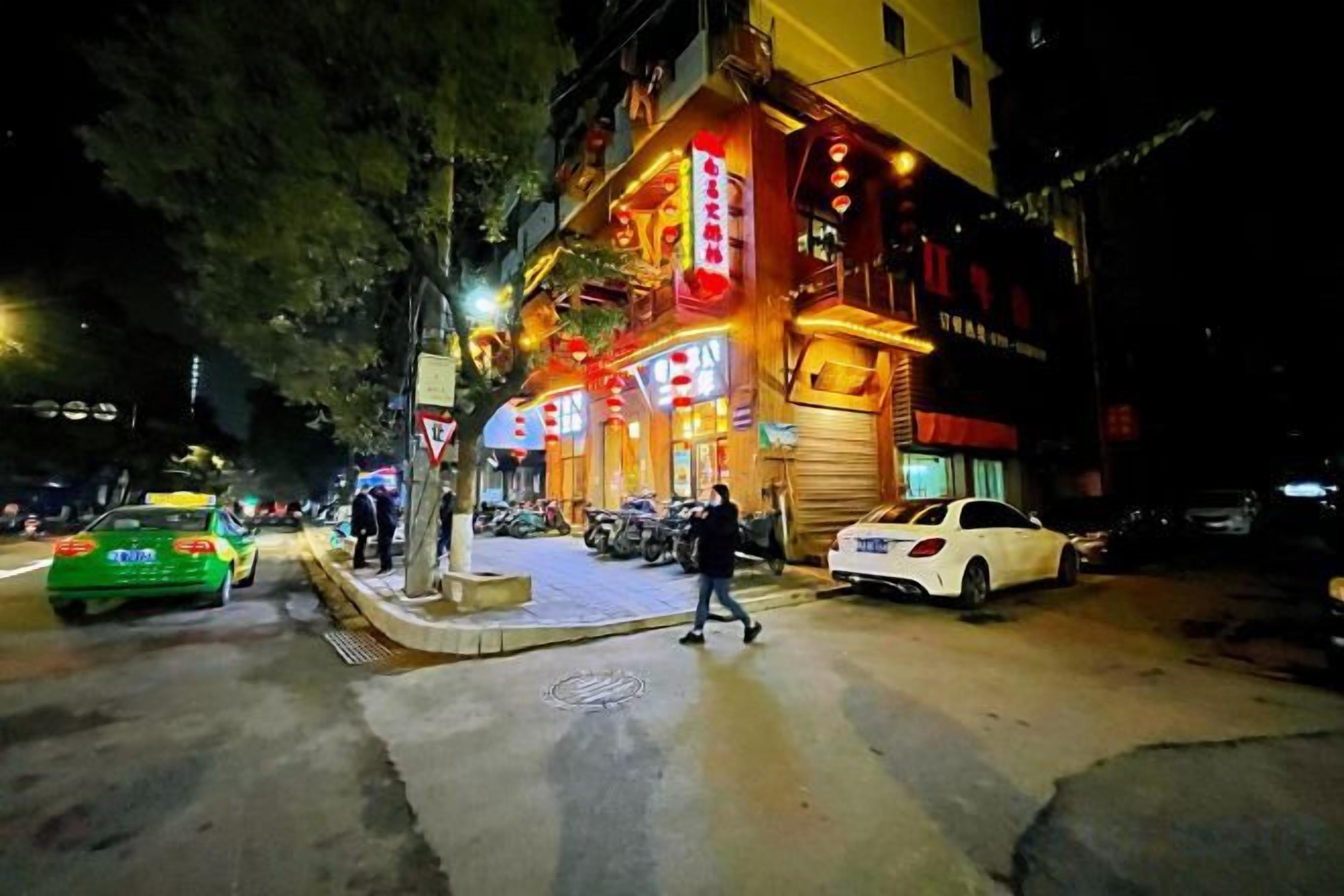 Una calle iluminada de noche con un edificio brillante, aceras y coches estacionados.
