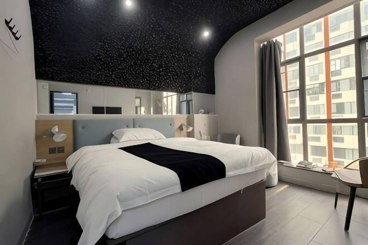 Un dormitorio moderno con una cama grande, una mesita de noche, una silla, una ventana con cortinas y una estantería en la pared con un espejo y un reloj.