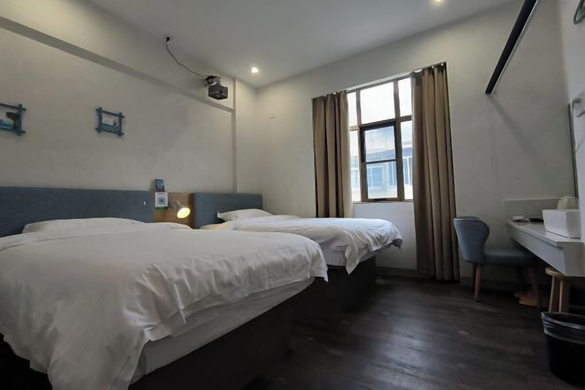 Ein Hotelzimmer mit zwei Betten, einem Schreibtisch, einem Stuhl, einem Fenster mit Vorhängen und einem an der Wand montierten Projektor.