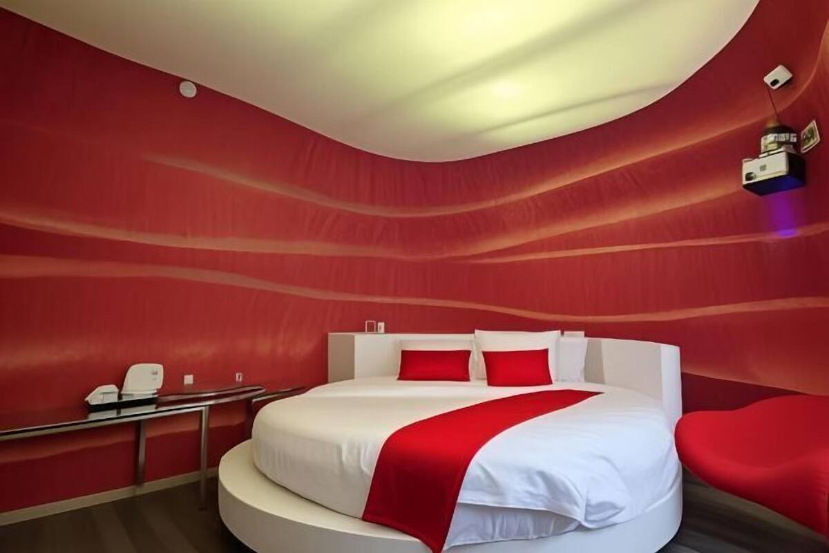 Una habitación de hotel moderna con una cama curva, paredes rojas y un escritorio pequeño con teléfono.