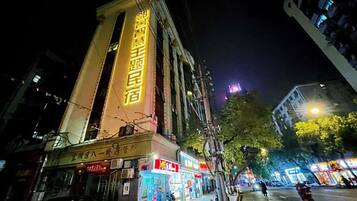 A building with illuminated signage, a street with shops, and a tree-lined sidewalk.