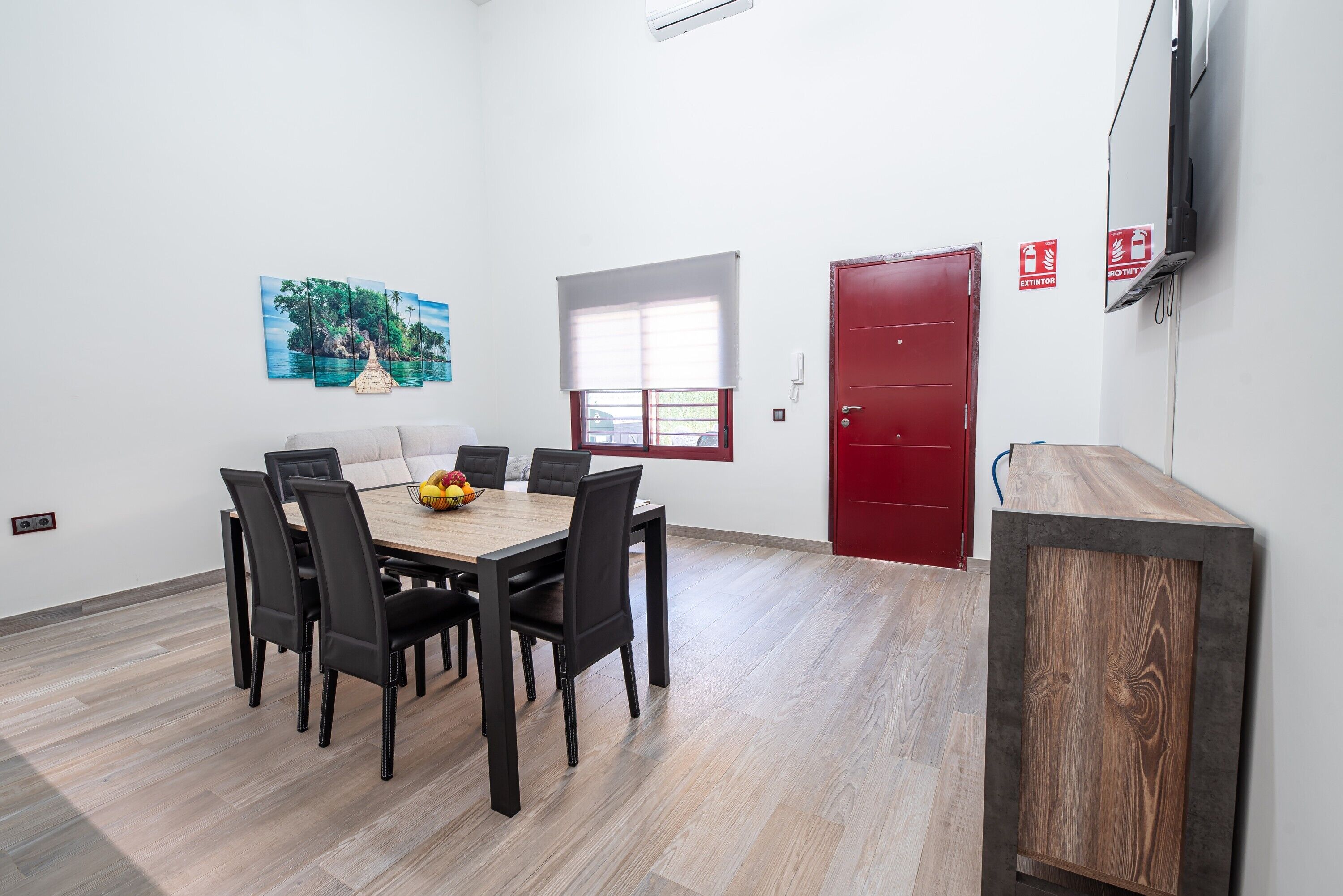 Un comedor con una mesa y sillas de madera, un cuadro en la pared, una ventana con persianas y una puerta roja.