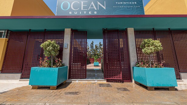 Entrada de un hotel con un letrero que dice 'OCEAN SUITES', diseño moderno y fachada azul y amarilla, y dos grandes macetas con plantas.