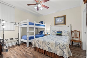 A room with bunk beds, a desk, a chair, a ceiling fan, and a framed picture on the wall.