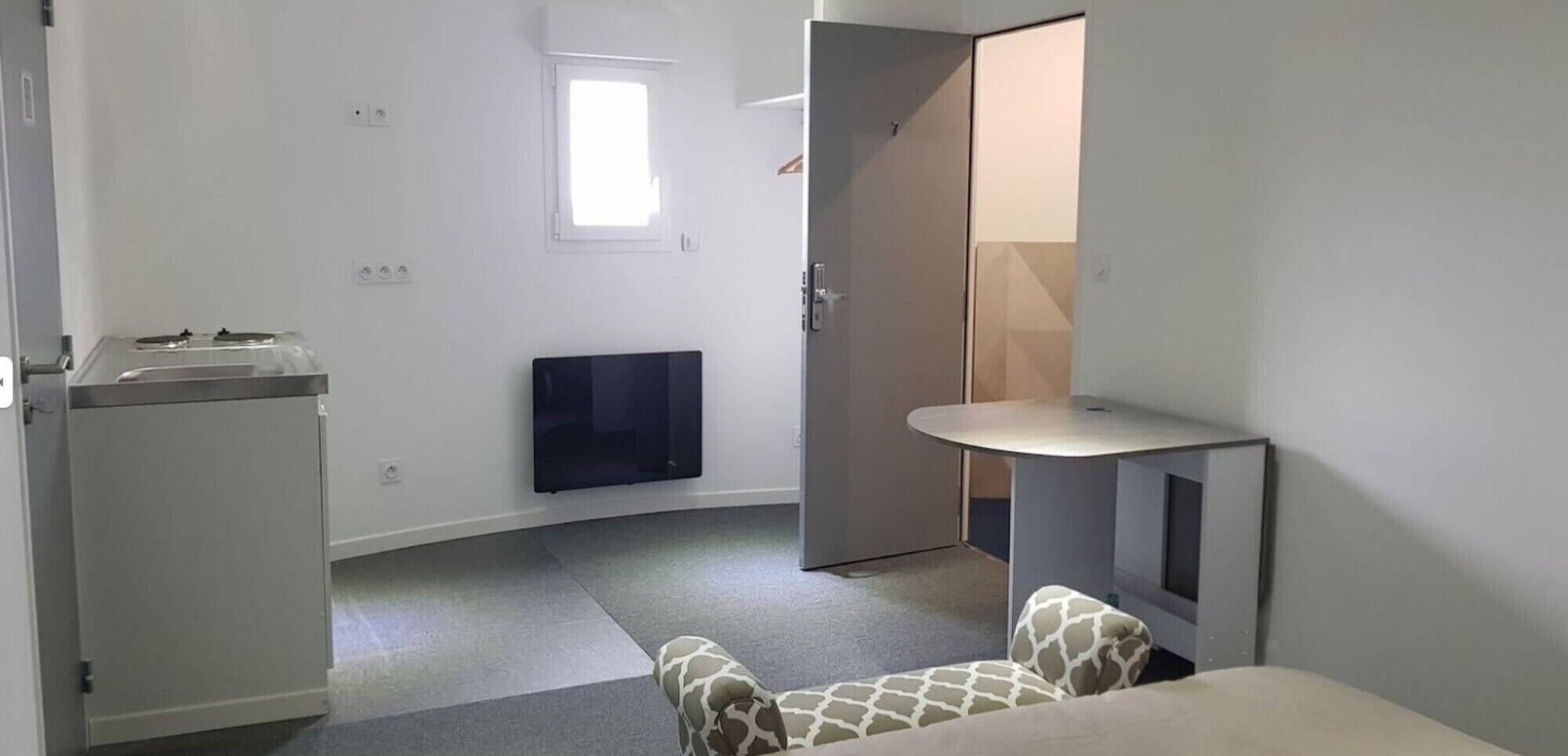 Une petite pièce compacte dotée d’une kitchenette, d’un téléviseur à écran plat fixé au mur, d’une petite table et d’une chaise à motifs.