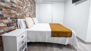 Una camera da letto con una parete in mattoni, un letto con biancheria da letto bianca e una coperta marrone, un comodino bianco con cassetti e una TV a parete.