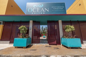 Entrada de un hotel con un letrero que dice 'OCEAN SUITES', diseño moderno y fachada azul y amarilla, y dos grandes macetas con plantas.