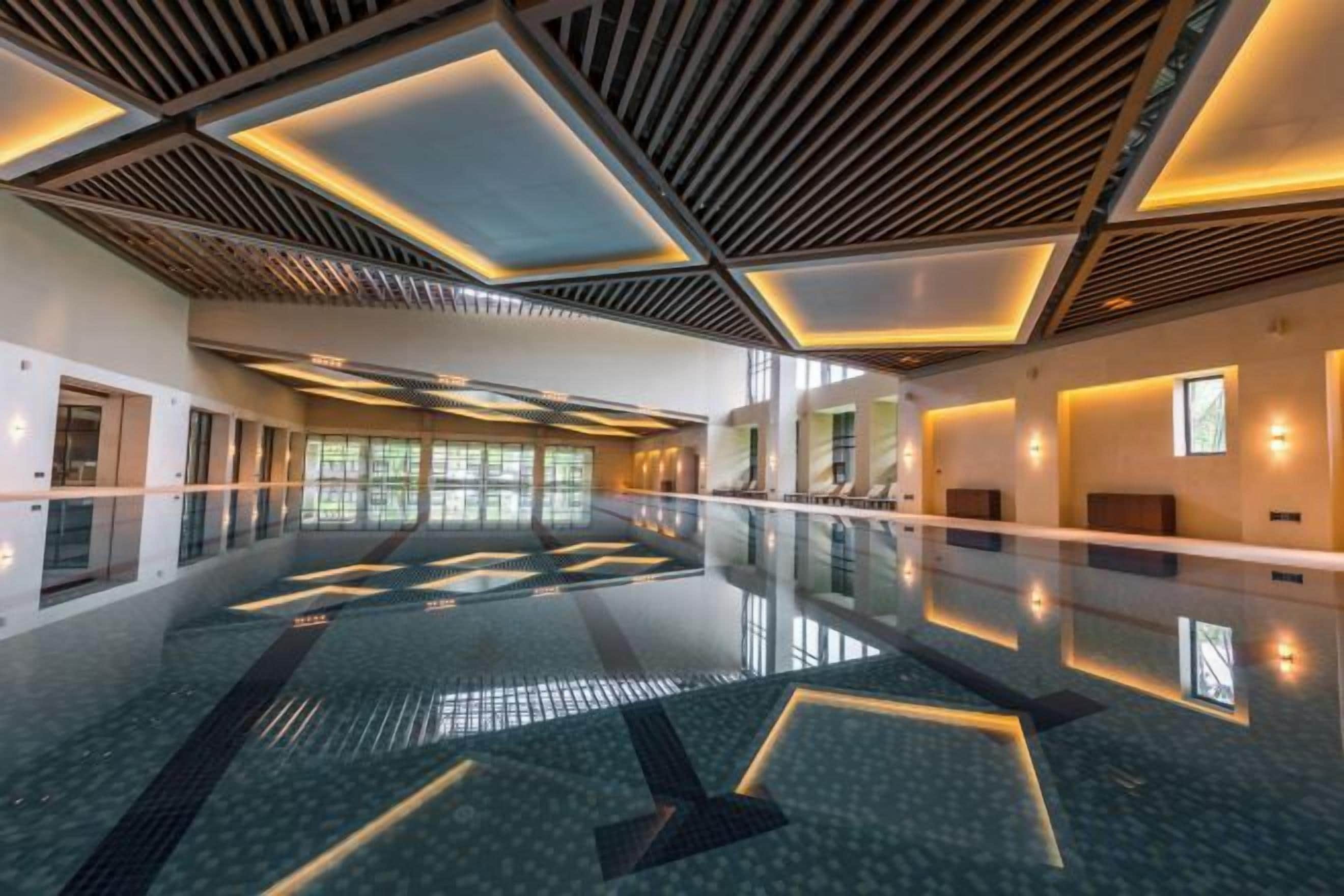Une vaste piscine intérieure au design moderne, avec des motifs géométriques sur le sol et le plafond, et un long couloir avec des fenêtres d’un seul côté.